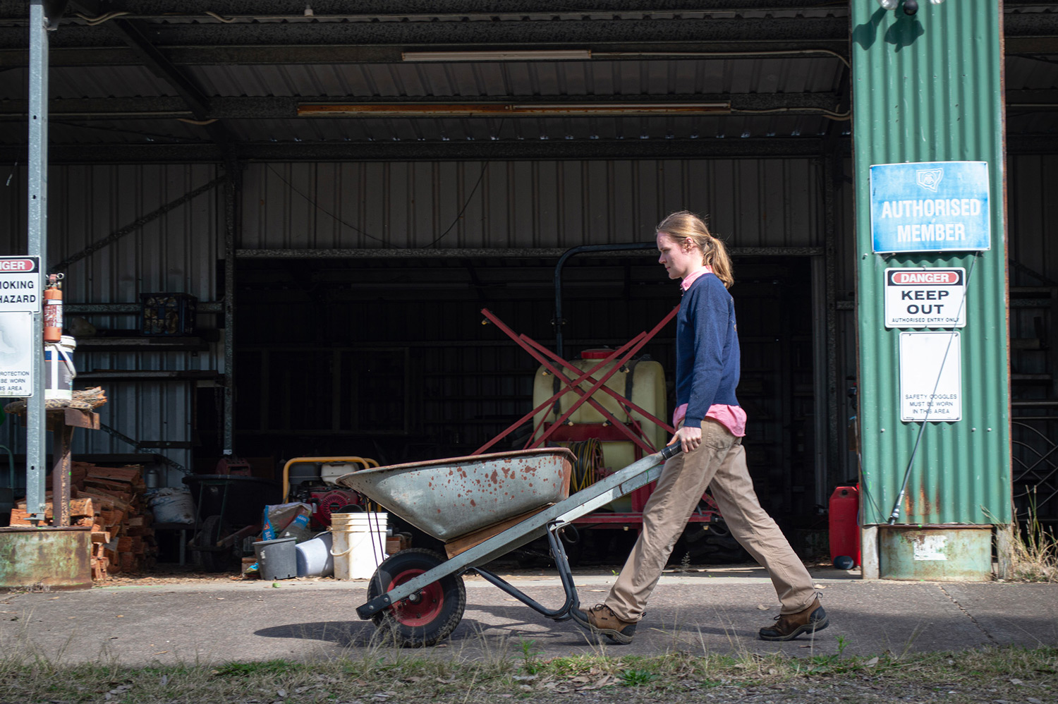 A woman pushes a wheelbarrow in front of a shed in a farm setting.