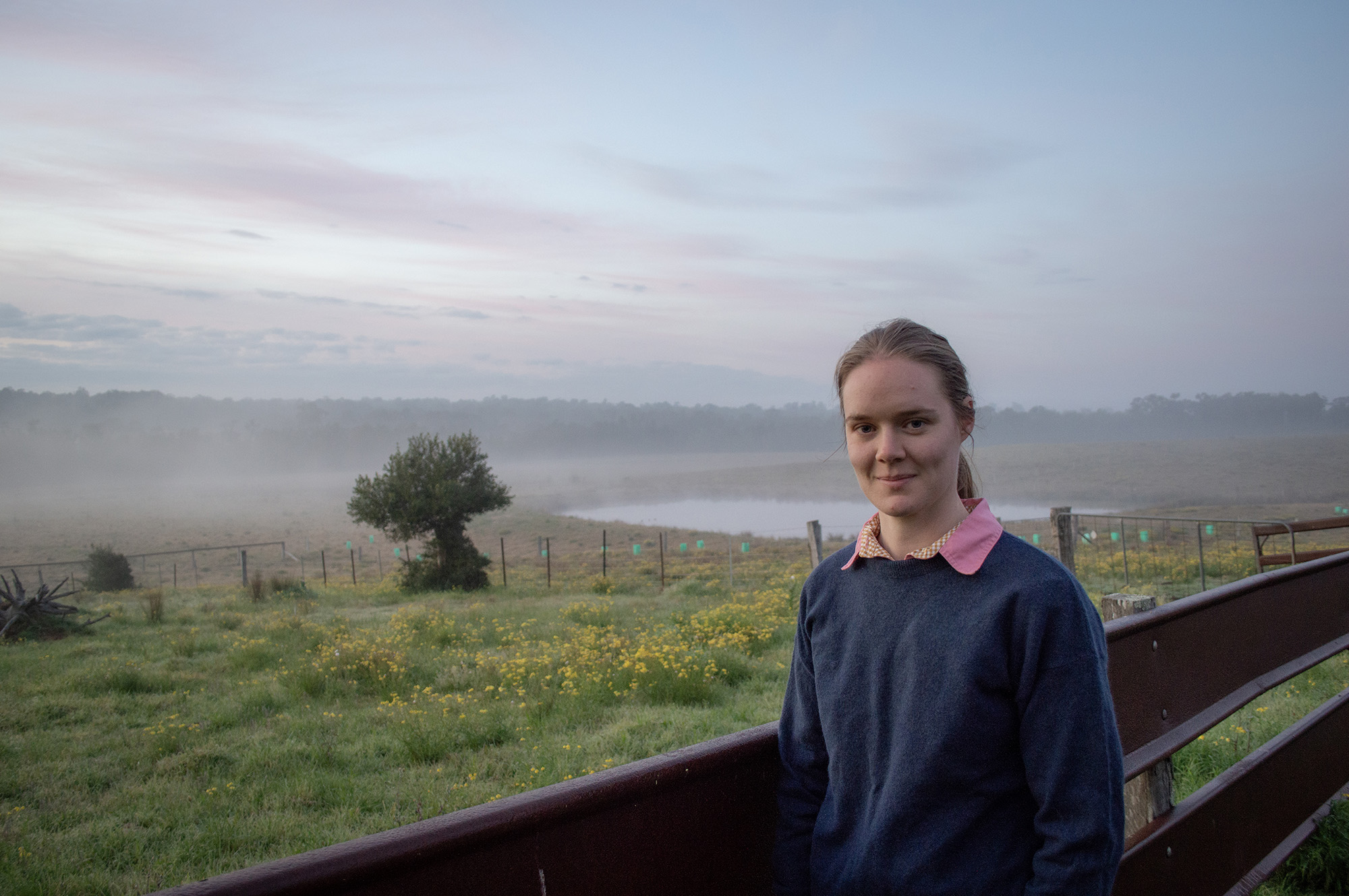 A woman stands in front of a paddock with flowers and a dam on a misty morning.