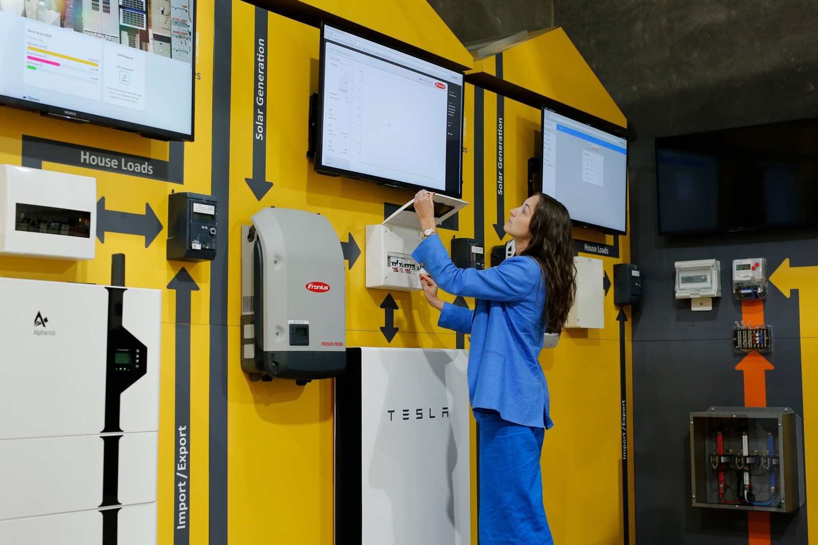 A woman dressed in a blue suit poses in front of a wall filled with several monitors, indicating a high-tech setting.