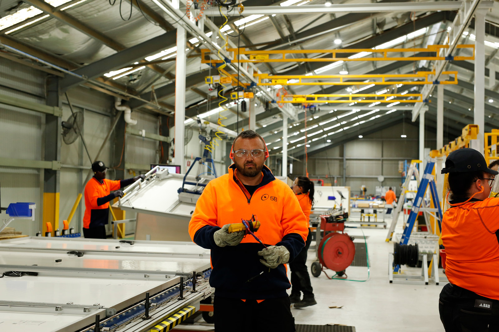 A man in hi-vis stands on an assembly line in a warehouse at a solar panel manufacturing plant.